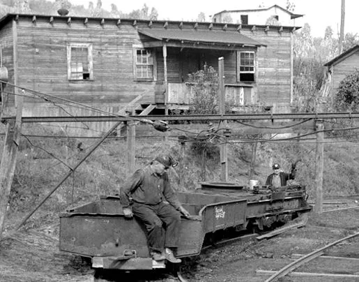 Early 20th century West Virginian miners sit on mine carts outside of a wooden house.