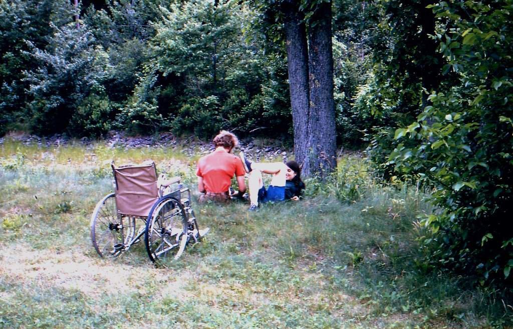 two camp members sit under a tree while attending a disabled persons camp; a wheel chair is focal point of image