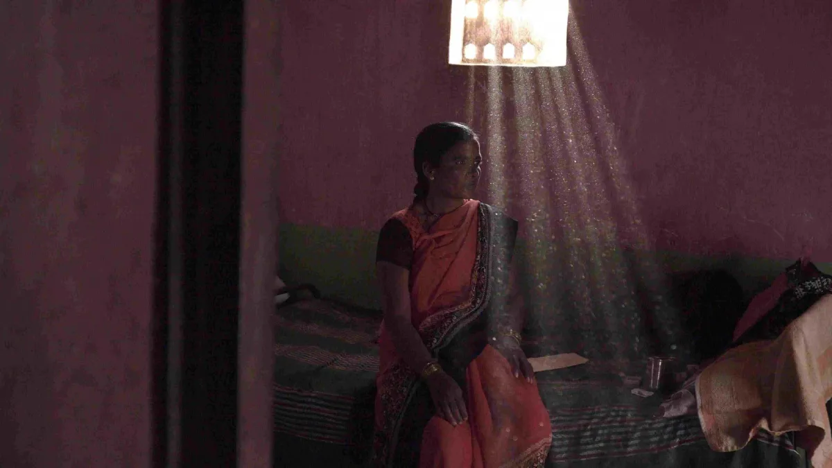 An Indian woman in a sari sits on a couch before a window. Light rays stream through the window's elaborate design