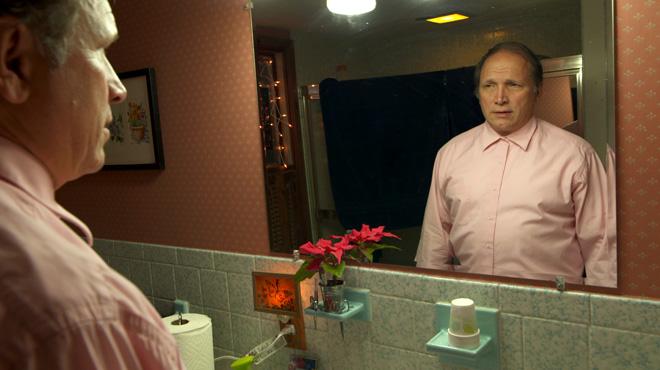 white middle aged male stares at himself in the bathroom mirror wearing pink button up