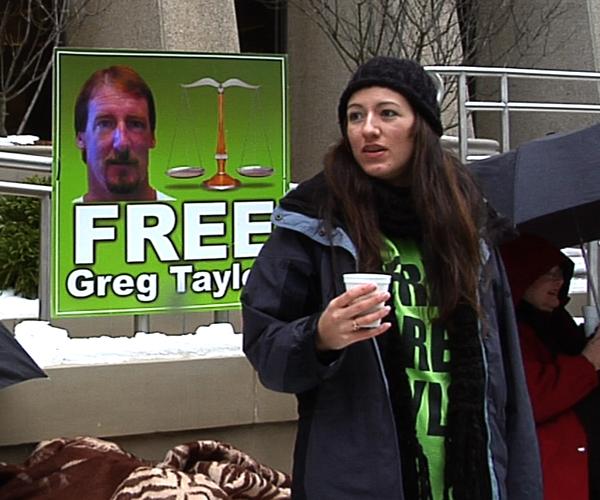 A woman stands in front of a 'Free Greg Taylor' poster.