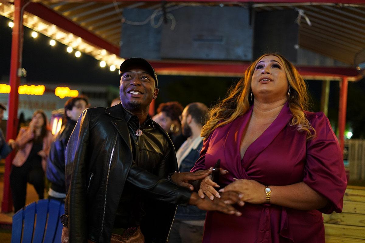 New Orleans city council candidate Mariah Moore, a Black Trans woman, wears a burgundy dress. She smiles and holds her brother's hand in anticipation of the incoming election results. 
