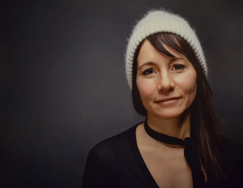 Headshot of a Latina woman with olive skin an long brown hair wearing all black and an off-white cashmere beanie. 