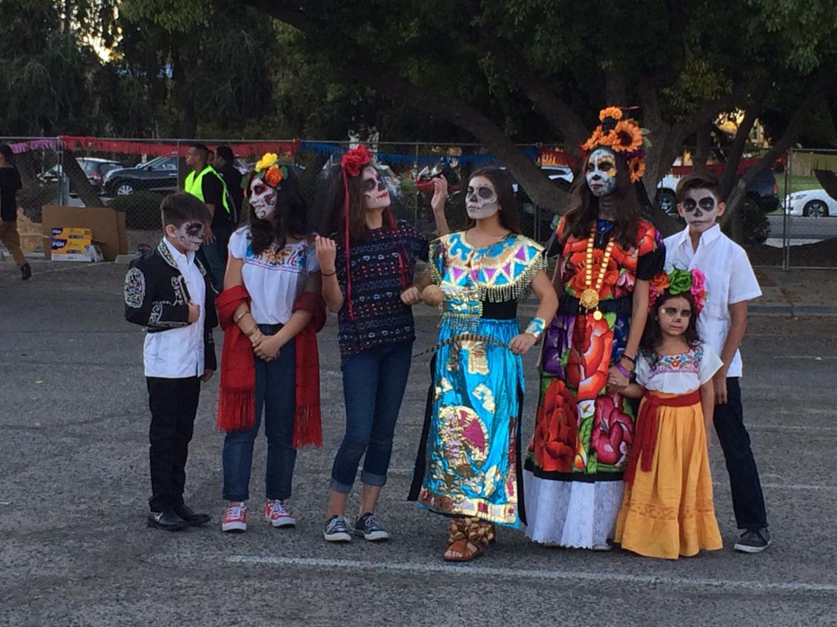 A group of Mexican-American women and children in Oaxacan dress and Dia de los Muertos make-up stand in a parking lot.