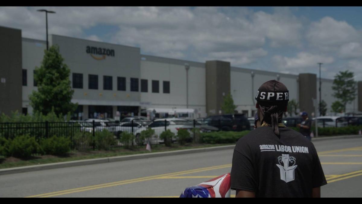 A man wearing an "Amazon Labor Union" t-shirt looking towards an Amazon Fulfillment Center warehouse. 