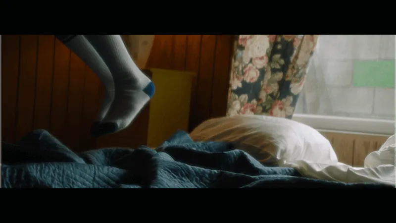 A pair of children's feet in white socks are visible jumping mid-air from a bed with a blue blanket