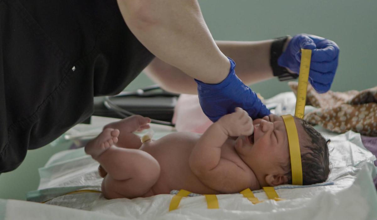 A pair of hands wearing medical gloves cares for a crying newborn infant. The nurse is using a tape measure to measure the circumference of the baby's head.
