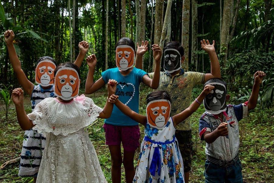 6 young children, in two rows of three, stand with their hands awkwardly raised in celebration. Behind them is a lush, green forest with tall and slim trees. The children are all wearing a paper mask that is a simple drawing of a young Hoolock Gibbon called Twiki. It is Twik's first birthday although he is not in the picture. We assume he is somewhere up in the trees behind them.