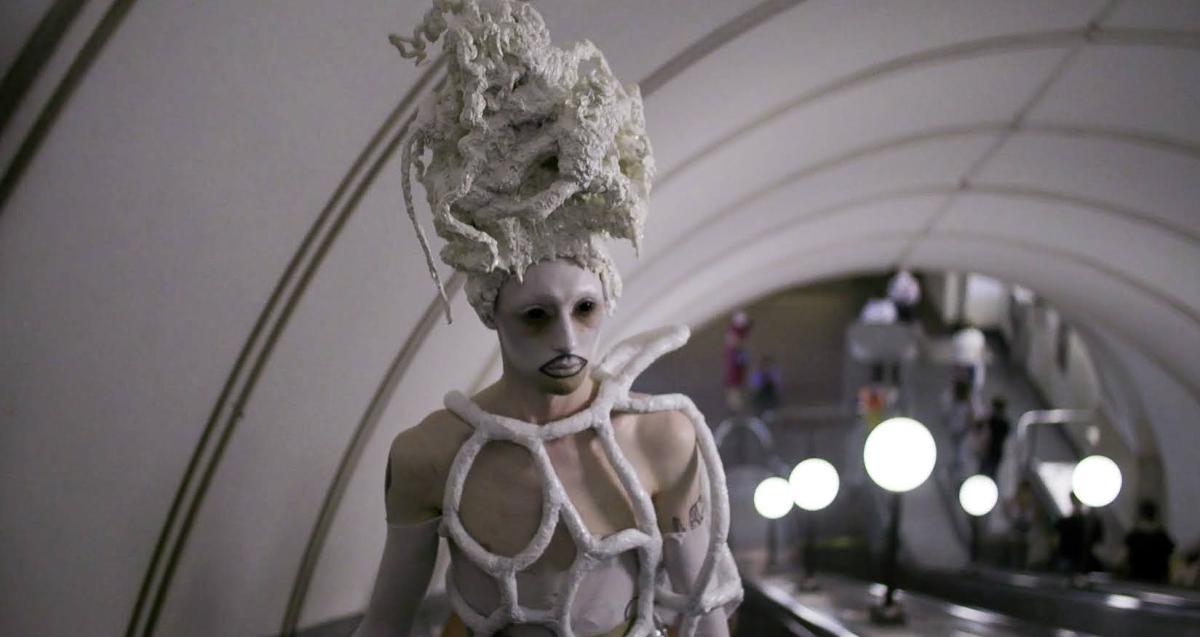Young person, wearing white makeup, black contact lenses, white outfit made of tubes and a tall hand-made headdress, is taking an escalator in subway.