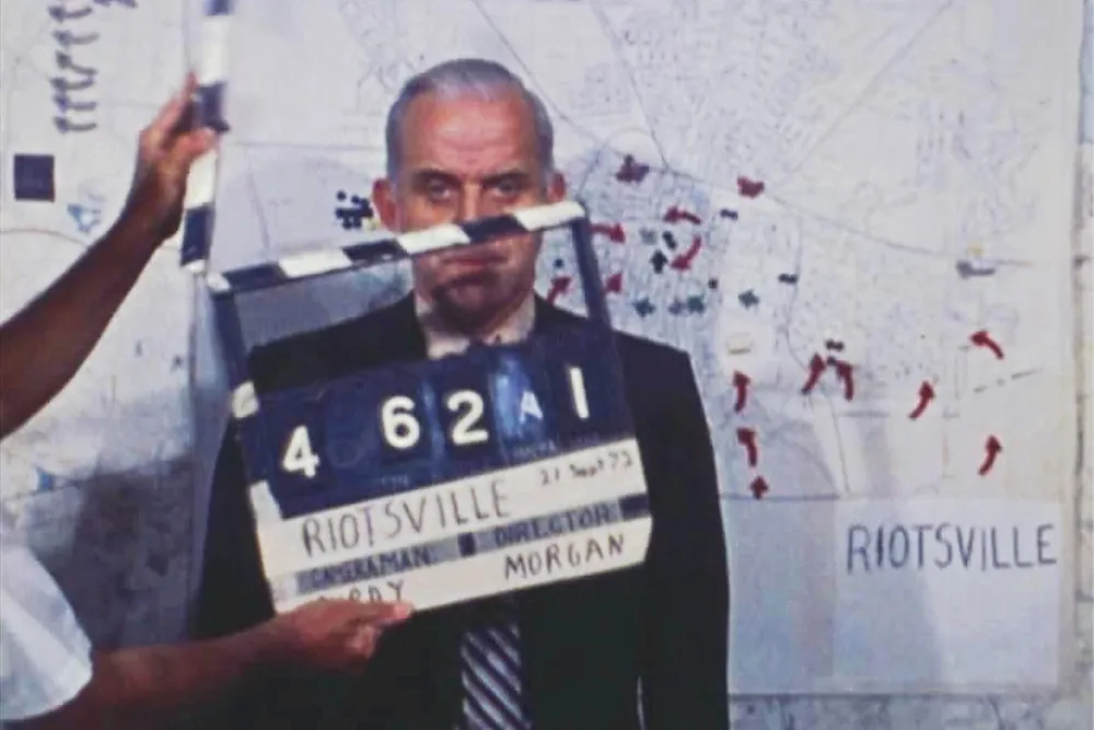 white male standing behind clapper board and in front of map labeled Riotsville with marked locations 