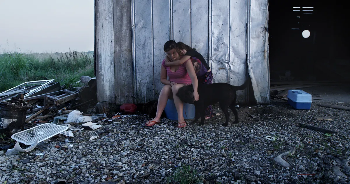 two female siblings sit on a plastic box and pet their black dog in front of a rural dilapidated barn.