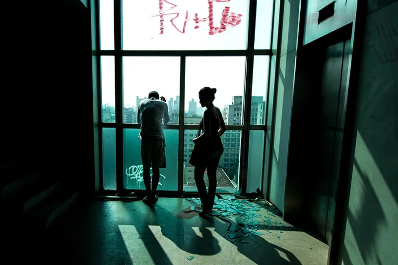 2 people inside a high-rise, standing next to an elevator. The window's glass is shattered.