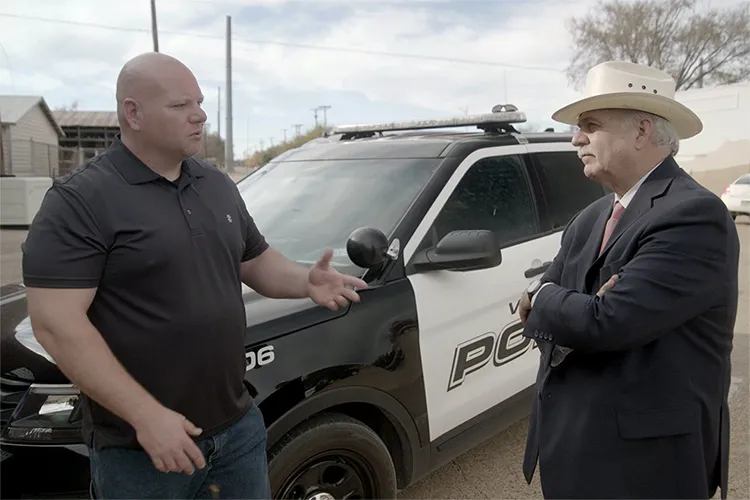 A man in a black shirt and a man in a suit and hat talk in front of a police cruiser.