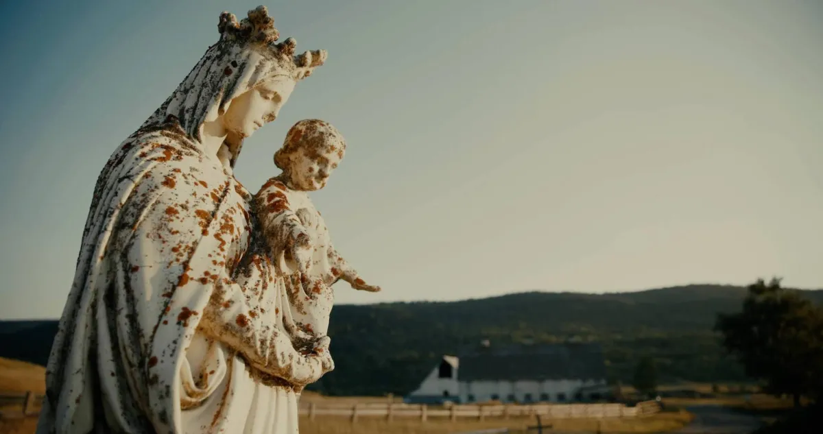 Large, white stone figure of Mary and baby Jesus in the foreground with barn and hills in the background. 