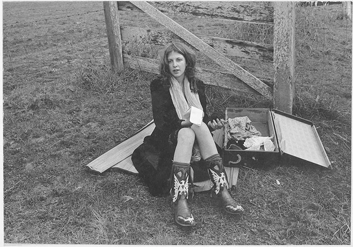 Young woman in early 70's fashion sits on a picnic blanket next to an open suitcase with a notepad in her hand.