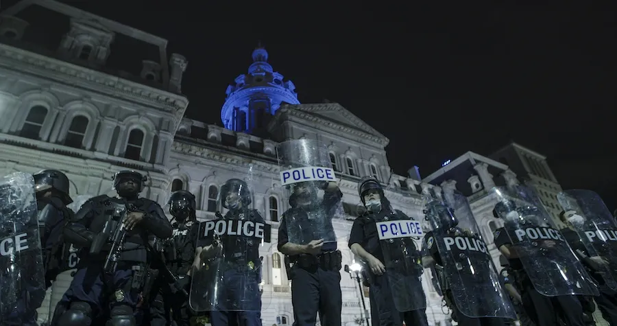 A line of Baltimore police officers wearing riot gear and holding shields stand in front of city hall the night after the murder of George Floyd.
