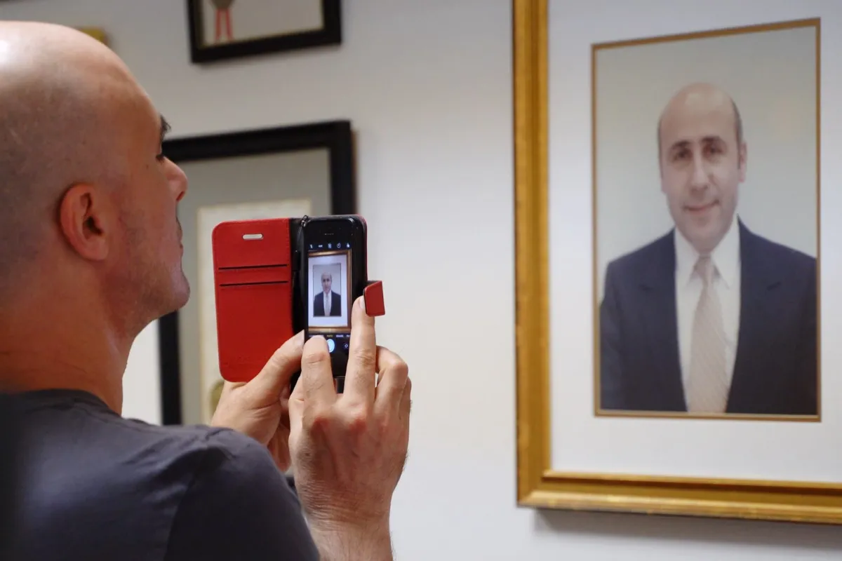 Bald white man takes a photo with an iPhone of a framed portrait of Alex Odeh. 
