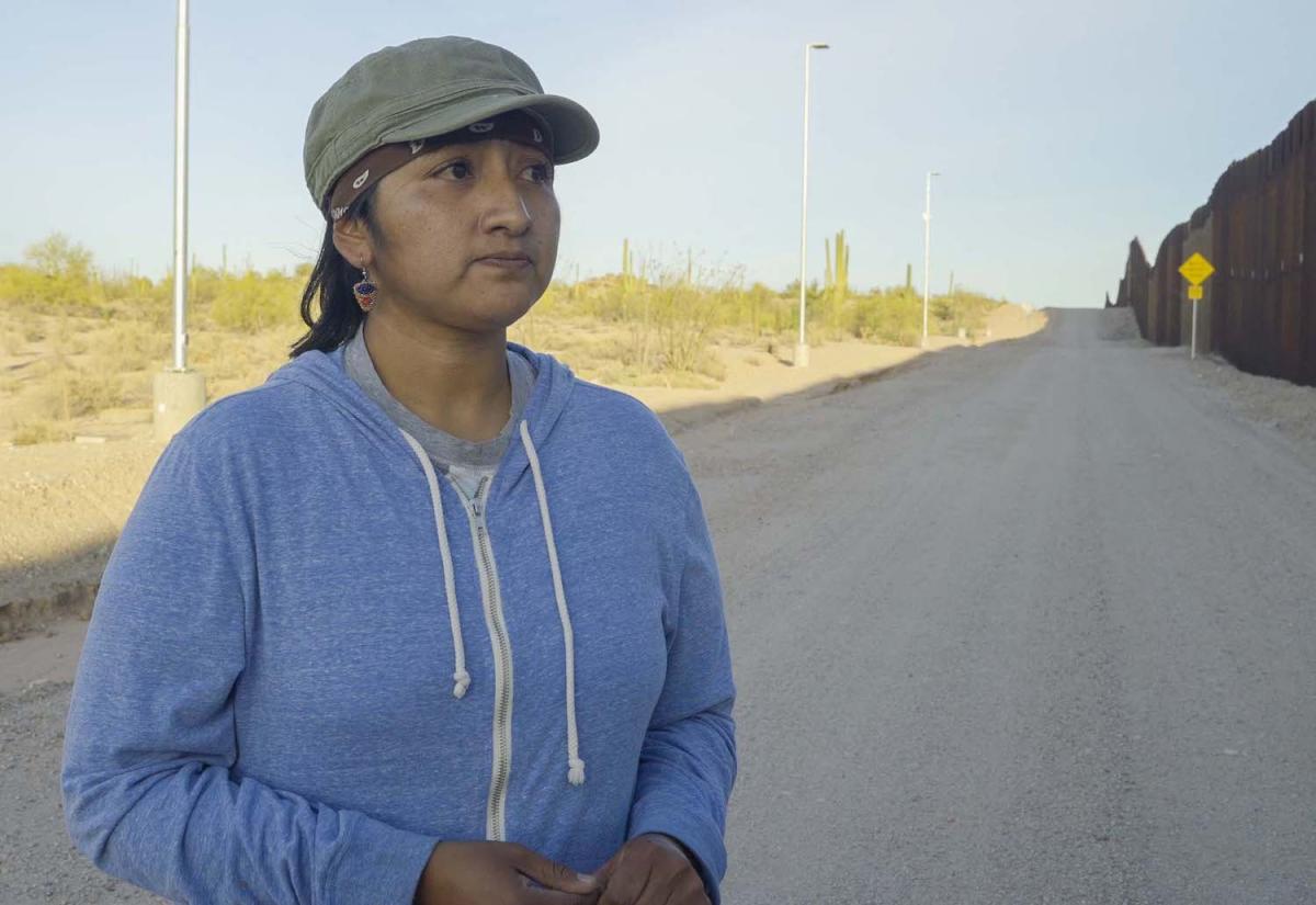 An indigenous woman stands in the shadow of the US-Mexico Border Wall