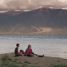Two women with short hair, and a dog, sit in front of a lake and mountains