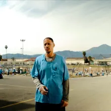 Man in blue prison shirt with a long sleeve olive green shirt underneath. Man has short brown hair. Standing in a prison yard.