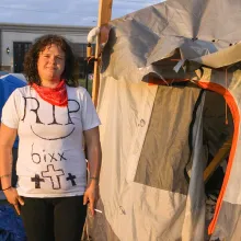 A homeless woman wearing a RIP 6ixx shirt stands next to an encampment