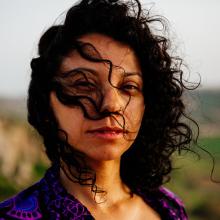 A woman with brown skin stares at the camera. Her dark brown curly hair drapes over her face, and a black and purple shawl is wrapped around her shoulders.