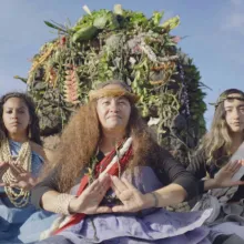 three women in authentic garb - light blue, light purple, and tan dress with sashes. They sit in front of an altar made of rock and other natural material