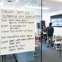 Conference room with yellow chairs, camera equipment and two people in the background wearing dark blue tops and gray bottoms. Sign on glass door in foreground with Conference Room schedule. 