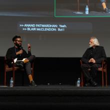 two people sitting in chairs on a stage. one person, dark brown skin, black glasses, black hair, dressed in all black, the other is fair skin complexion, with white hair, dress in all black