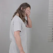 A Caucasian man with long hair covers his face with his hand while standing in front of a mugshot background 