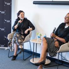 Two people sitting in chairs. Person on left is holding a microphone up to their face, wearing a floral skirt, black sweater, and black shoes. Person on right is holding a microphone down with a big smile, wearing a black and white polka dot skirt with a black sweater and black heels.