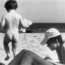 Black and white photo of a naked baby running on the beach. Adult person is laying in the sand on stomach wearing a baseball cap, swimsuit, and sunglasses.