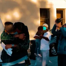 Images courtesy of "What These Walls Won't Hold" film: Under dappled sunlight in a parking lot, a family reunifies with a loved one. Standing a respectful distance away is a photographer wearing a blue puffy jacket