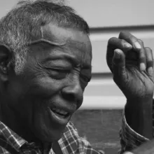 A black & white image of an elderly man of African-American descent sitting on a porch.