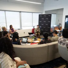 Photograph of a room filled with multiple people seated and clapping in applause. The focus is on Chris Boeckmann, Getting Real '22 Programmer, who is wearing brown pants and a light olive green long-sleeved button-up, sitting in front of a black backdrop.