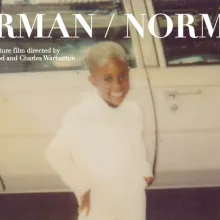 Photograph of a Black child dressed in white standing in front of an older white car, the text Norman/Norman at the top of the image