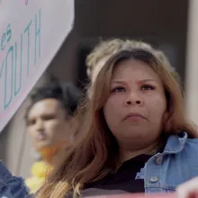 A young woman stands in a crowd of people, holding a sign that says "Protect Futures of Youth". 