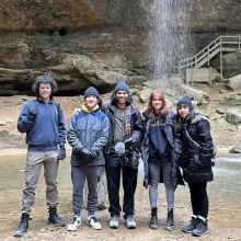 Five donor-conceived siblings wearing winter clothes stand in front of a waterfall. 