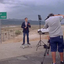 A news anchor stands in the middle of the desert, in front of a sign that says, “ELY STATE PRISON”. A production light shines on them, and a camera person with a tripod films them. 