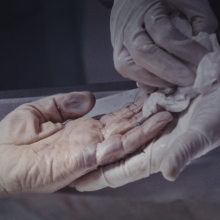 Still image from 'Pure Unknown,' depicting a pair of gloved hands cradling and gently wiping the hand of someone who is deceased.