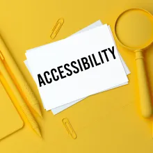 Graphic with yellow background featuring a yellow pen, yellow pencils, yellow paperclips, a yellow magnify glass, a yellow cellphone, and a stack of white note cards. The top note card has the word Accessibility written in black. 