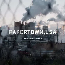 A factory seen from behind a wire fence, the text "Papertown, USA - A Documentary Film" is superimposed on the image in white 