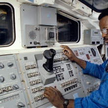 Still is from DC/DOX's Closing Night film 'The Space Race.' A Black man wearing glasses and a blue space suit with the American flag patch on the left arm, stands in front of a console on a space craft. 