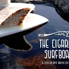 The tail of a surfboard made out of cigarette butts floats on water, the text "The Cigarette Surfboard: A Film by Ben Judkins" is written in the bottom right corner in white