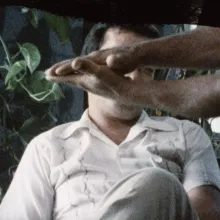 A man sits in a beige button up in a chair in front of greenery. In front of him hands and arms clasping, as if they are slating on a movie set.