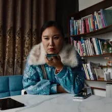 A Chinese woman with dark hair and a sequined blue jacket with a fur collar sits at a white marble table and speaks into her cell phone. Another cell phone rests on the table by her elbow. There is a brown curtain and wooden bookshelf behind her.