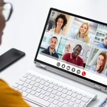 A person wearing a gold shirt is looking at a laptop with six faces on the screen having a virtual meeting. 