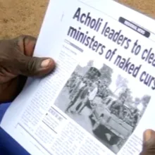 Image of two hands holding a folded newspaper with the headline "Acholi leaders to cleanse ministers of naked curse"