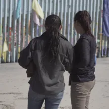 Photographed from behind, two people with long black hair stand in front of a tall, grey border wall.