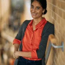 Person standing on stairs, leaning against a brown railing and wall. Person is wearing an burnt orange button up, black pants, and a dark gray cardigan. Their dark hair is in a low bun to the left side. 